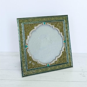 May include: A square picture frame featuring an olive green and gold metallic finish with intricate embossed floral patterns. The central opening is scalloped and bordered by silver-toned metal accents, embellished with small turquoise and amber rhinestones.