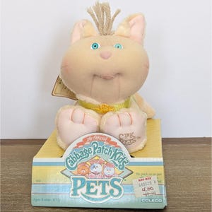 May include: A beige plush cat toy with blue eyes, a yellow collar, and a tuft of yarn hair on its head. The toy sits on a yellow and blue cardboard display box labeled Cabbage Patch Kids Pets with a Kay-Bee price sticker on the front.