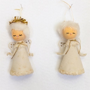 May include: Two hanging angel ornaments featuring white felt cone-shaped bodies, lace trim, and sheer wings with gold accents. Each figure has a round peach-colored face, closed eyes, a small pink nose, and white hair, with one wearing a gold tinsel halo.