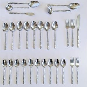 May include: A collection of stainless steel silverware with bamboo-style handles. The set includes spoons of various sizes, forks, and knives. The silverware is arranged on a white surface, showcasing its polished, reflective surfaces.