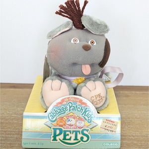 May include: A grey plush Cabbage Patch Kids pet toy with brown yarn hair, a pink tongue, and light pink paws. The toy features embroidered CPK PETS text on one paw and sits in a yellow and blue cardboard display box with the Cabbage Patch Kids logo.