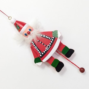 May include: A wooden Santa Claus jumping jack ornament featuring a red hat with holly, a white faux fur beard, and a red coat with black and white trim. The figure has articulated limbs connected by a red string with a small wooden bead at the end.