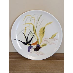 May include: White ceramic plate with a painted botanical design. The design features black bird silhouettes, yellow and green foliage, purple grapes, and orange and brown accents. The plate is round and sits on a wooden surface.