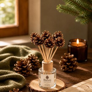 May include: A clear glass reed diffuser bottle with a light wood cap, featuring a white label with a pinecone icon and the text BiZen REED DIFFUSER HANDMADE COLORADO. Several thin wooden reeds topped with small brown pinecones emerge from the bottle.