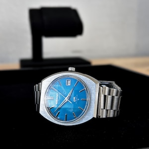 May include: A silver-toned Seiko automatic wristwatch featuring a vibrant blue dial with silver hour markers and hands. The watch includes a date window at the three o'clock position, the text Seiko Automatic and LM 17 Jewels on the face, and a brushed metal link bracelet.