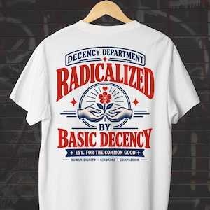 May include: A white t-shirt featuring a graphic on the back with the text "DECENCY DEPARTMENT RADICALIZED BY BASIC DECENCY EST. FOR THE COMMON GOOD HUMAN DIGNITY KINDNESS COMPASSION" in red and blue, with an illustration of hands holding a flower.