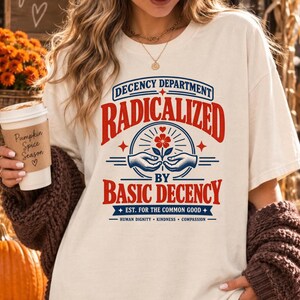 May include: A cream-colored t-shirt featuring red and blue text that reads Decency Department Radicalized By Basic Decency Est. For The Common Good Human Dignity Kindness Compassion, with a graphic of two hands holding a flower.