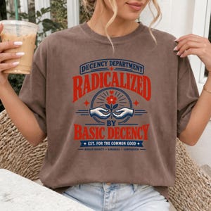 May include: A brown short-sleeve t-shirt featuring a graphic print with the text "DECENCY DEPARTMENT RADICALIZED BY BASIC DECENCY EST. FOR THE COMMON GOOD HUMAN DIGNITY KINDNESS COMPASSION" and an illustration of two hands holding a flower.