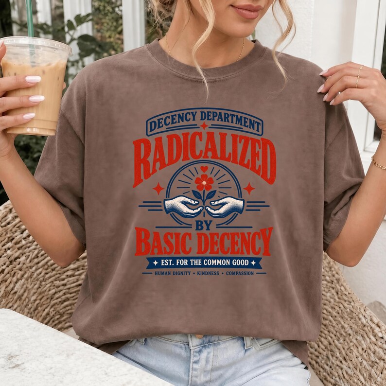 May include: A brown short-sleeve t-shirt featuring a graphic print with the text "DECENCY DEPARTMENT RADICALIZED BY BASIC DECENCY EST. FOR THE COMMON GOOD HUMAN DIGNITY KINDNESS COMPASSION" and an illustration of two hands holding a flower.