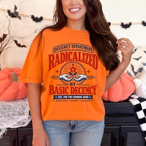 May include: An orange t-shirt featuring a graphic with the text "DECENCY DEPARTMENT RADICALIZED BY BASIC DECENCY EST. FOR THE COMMON GOOD HUMAN DIGNITY KINDNESS COMPASSION" and an illustration of two hands holding a flower in red and blue ink.