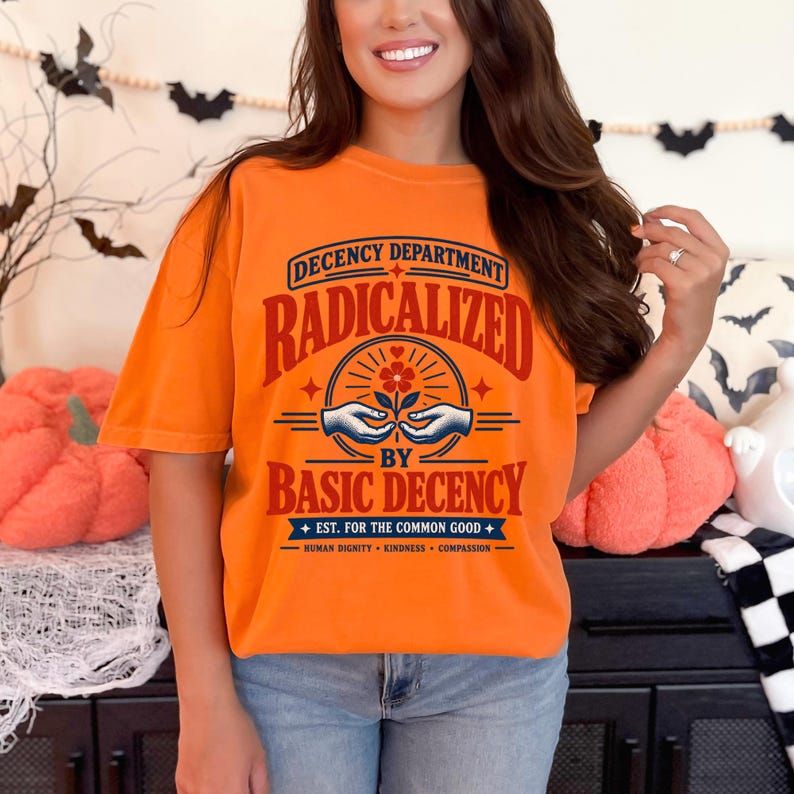 May include: An orange t-shirt featuring a graphic with the text "DECENCY DEPARTMENT RADICALIZED BY BASIC DECENCY EST. FOR THE COMMON GOOD HUMAN DIGNITY KINDNESS COMPASSION" and an illustration of two hands holding a flower in red and blue ink.