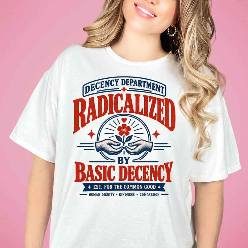 May include: A white t-shirt featuring a graphic design with red and blue text that reads "DECENCY DEPARTMENT RADICALIZED BY BASIC DECENCY EST. FOR THE COMMON GOOD HUMAN DIGNITY KINDNESS COMPASSION" and an illustration of hands holding a flower.