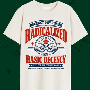 May include: A cream-colored short-sleeve t-shirt featuring a graphic print in red and blue. The text reads: DECENCY DEPARTMENT RADICALIZED BY BASIC DECENCY EST. FOR THE COMMON GOOD HUMAN DIGNITY KINDNESS COMPASSION, with an illustration of hands.