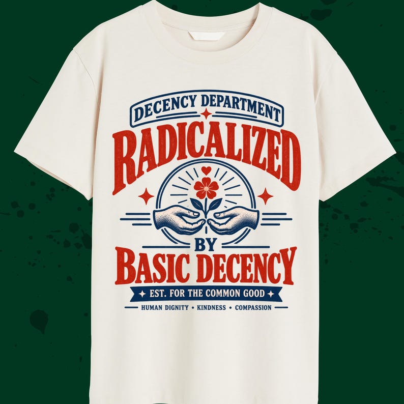 May include: A cream-colored short-sleeve t-shirt featuring a graphic print in red and blue. The text reads: DECENCY DEPARTMENT RADICALIZED BY BASIC DECENCY EST. FOR THE COMMON GOOD HUMAN DIGNITY KINDNESS COMPASSION, with an illustration of hands.
