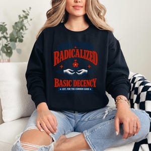 May include: A black long-sleeve sweatshirt featuring a graphic print in red and blue. The text reads RADICALIZED BASIC DECENCY and EST. FOR THE COMMON GOOD, accompanied by an illustration of two hands holding a small red flower.