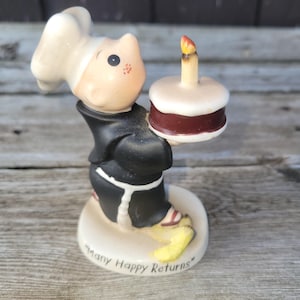 May include: A ceramic figurine of a cat-like character in a black robe and chef's hat, holding a small cake with a lit candle. The base reads "Many Happy Returns". The figurine is painted with details in white, red, and yellow.