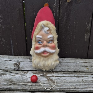 May include: A vintage Santa Claus head ornament featuring a red fabric hat and a cream-colored faux fur beard and trim. The face has painted blue eyes, rosy cheeks, and a white mustache. A thin white string with a small red ball hangs from the bottom.