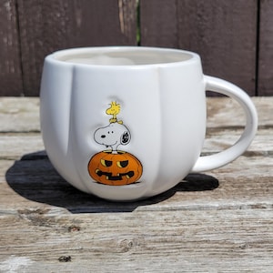 May include: White ceramic mug with a pumpkin shape and a handle. The mug features a cartoon illustration of Snoopy and Woodstock on a jack-o'-lantern. The mug is perfect for enjoying hot beverages during the Halloween season.