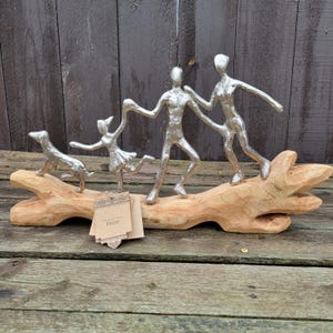 May include: A decorative sculpture featuring a family and a dog, all in silver-colored metal, atop a natural wood base. The figures are stylized, with outstretched arms, suggesting movement. The wood base has a natural, light brown color. A tag with the word "INDE" is attached.