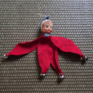 May include: A vintage-style elf ornament featuring a painted face with blue eyes and rosy cheeks. The figure wears a grey felt hat and a matching grey bowtie. Its body consists of four red felt petal-like appendages, each tipped with a small silver bell.