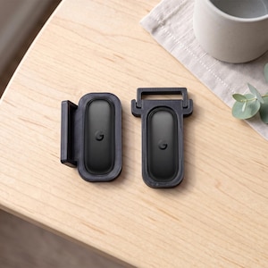 May include: Two black oval-shaped electronic devices with a small letter G logo in the centre. Each device is housed in a black plastic frame, with one frame featuring a side-mounted bracket and the other featuring a top-mounted rectangular loop.