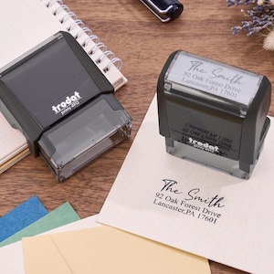 Op de afbeelding: Twee zwarte Trodat Printy 4912 zelfinktende stempels. Eén stempel is gesloten, terwijl de andere zo is geplaatst dat een afdruk op een witte envelop zichtbaar is: The Smith, 92 Oak Forest Drive, Lancaster, PA 17601 in een script en serif lettertype.
