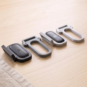 May include: Four small, rectangular, hinged plastic stands arranged in a row. Two stands are black and grey with an oval black insert featuring a small circular logo, while the other two are light grey and dark grey frames without inserts.