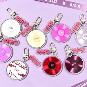 May include: A collection of circular keychains with various colors and designs. Each keychain features a disc-like charm with names like "SUGA", "J-hope", and "Jimin". The keychains are attached to silver clasps and connected by pink accents.