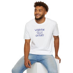 May include: A white short-sleeved crew neck t-shirt featuring the text WORK'EM SILLY, GATORS! printed in blue on the chest. The shirt is paired with light blue denim jeans, creating a casual look with a simple graphic design on the front.