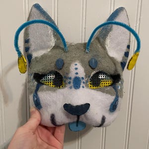 K&ouml;nnte beinhalten: Eine handgefertigte Katzenmaske aus Filz mit grauen, wei&szlig;en und blauen Akzenten. Die Maske hat blaue Pfeifenputzer-Ohren mit gelben Akzenten, schwarze Netzhautaugen und eine blaue Zunge. Das Wort "Front" ist sichtbar.