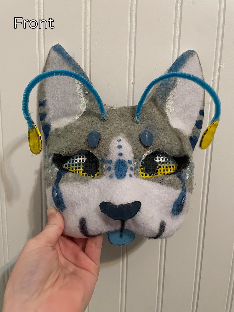 K&ouml;nnte beinhalten: Eine handgefertigte Katzenmaske aus Filz mit grauen, wei&szlig;en und blauen Akzenten. Die Maske hat blaue Pfeifenputzer-Ohren mit gelben Akzenten, schwarze Netzhautaugen und eine blaue Zunge. Das Wort "Front" ist sichtbar.