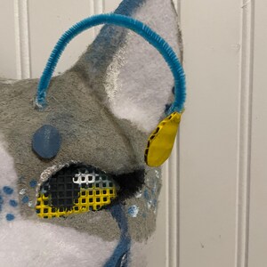 K&ouml;nnte beinhalten: Nahaufnahme einer grau-wei&szlig;en Fellmaske mit blauem Pfeifenreiniger und gelben Akzenten. Die Maske hat ein Netzauge und blaue Tr&auml;nendetails. Das Wort "Closeup" ist in der oberen linken Ecke sichtbar.