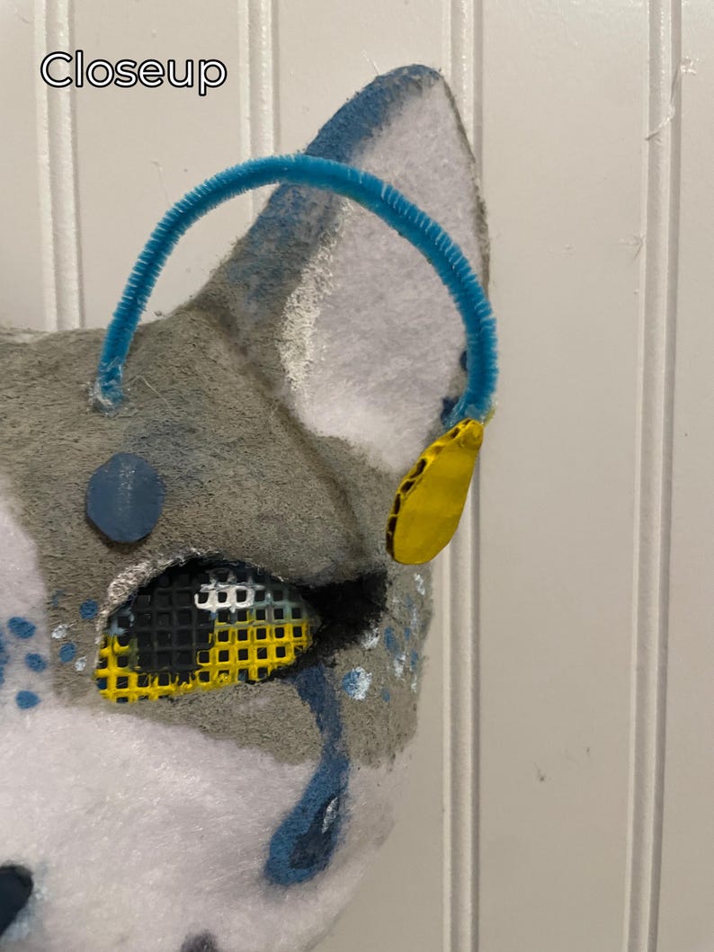K&ouml;nnte beinhalten: Nahaufnahme einer grau-wei&szlig;en Fellmaske mit blauem Pfeifenreiniger und gelben Akzenten. Die Maske hat ein Netzauge und blaue Tr&auml;nendetails. Das Wort "Closeup" ist in der oberen linken Ecke sichtbar.