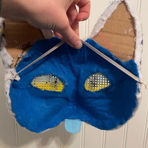 K&ouml;nnte beinhalten: R&uuml;ckansicht einer handgefertigten blau-wei&szlig;en Katzenmaske. Das Innere besteht aus blauem Filz mit Augen&ouml;ffnungen. Ohren aus Pappe und wei&szlig;em Stoff. Eine kleine blaue Zunge ist befestigt. Der Text "Back/Inside" ist sichtbar.