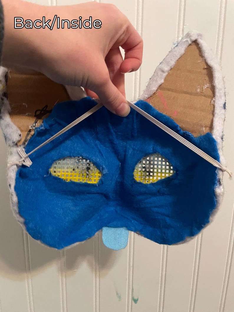 K&ouml;nnte beinhalten: R&uuml;ckansicht einer handgefertigten blau-wei&szlig;en Katzenmaske. Das Innere besteht aus blauem Filz mit Augen&ouml;ffnungen. Ohren aus Pappe und wei&szlig;em Stoff. Eine kleine blaue Zunge ist befestigt. Der Text "Back/Inside" ist sichtbar.