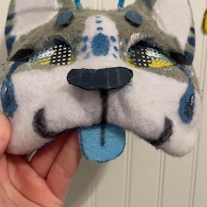 K&ouml;nnte beinhalten: Eine handgefertigte Hundemaske aus Filz mit einer blauen Zunge. Die Maske hat graues und wei&szlig;es Fell mit blauen Akzenten um Augen und Ohren. Der Text "Closeup" ist oben links sichtbar.