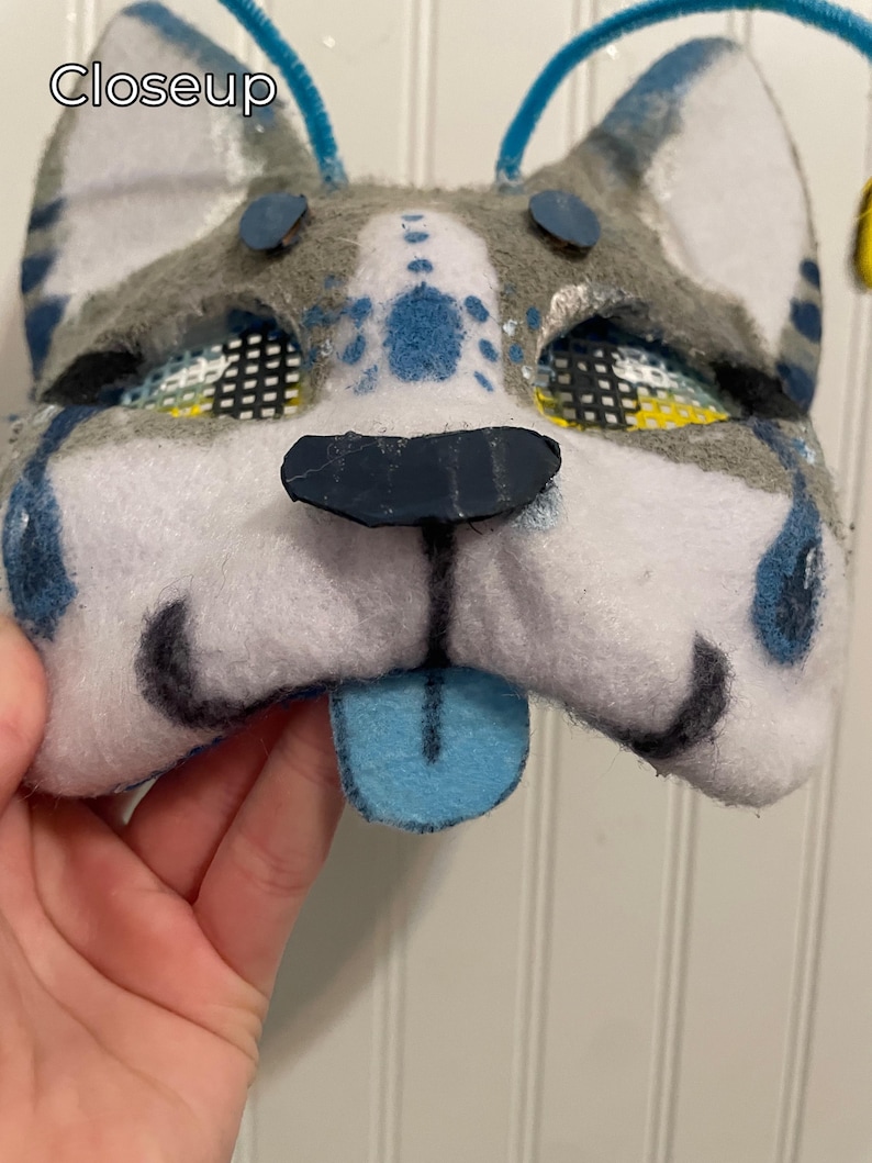 K&ouml;nnte beinhalten: Eine handgefertigte Hundemaske aus Filz mit einer blauen Zunge. Die Maske hat graues und wei&szlig;es Fell mit blauen Akzenten um Augen und Ohren. Der Text "Closeup" ist oben links sichtbar.