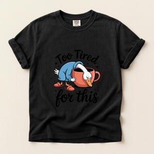 May include: Black t-shirt featuring a cartoon duck wearing a blue robe and slippers, leaning into a red mug. The text "Too Tired for this" is above and below the duck and mug illustration.