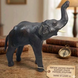 May include: A vintage leather-wrapped elephant figurine sculpture with its trunk raised. The dark gray elephant has ivory tusks and is labeled "Trunk Up Lucky 11". The sculpture is on a wooden surface.