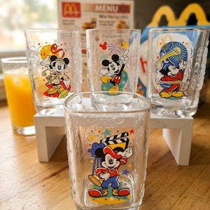May include: A set of four clear glass tumblers featuring colorful illustrations of Mickey Mouse in various outfits. Each glass displays a unique scene with stars and festive elements. The front glass shows Mickey with a movie clapperboard.