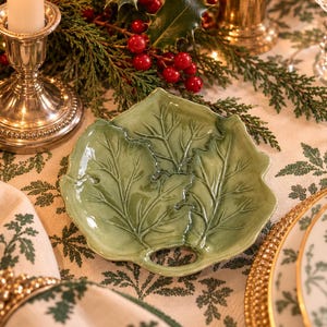May include: A light green ceramic dish shaped like a holly leaf with embossed veins and a small circular cutout at the base. The dish has a glossy finish and features a detailed leaf structure with pointed edges, centered on a patterned tablecloth.