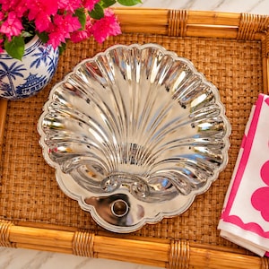 May include: A polished silver-toned metal dish shaped like a large scallop shell. The dish features a scalloped edge, deep radial grooves, and a circular opening at the base. The reflective surface shows light highlights and shadows.