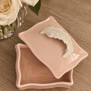May include: A rectangular ceramic trinket box with a glossy light pink finish and wavy edges. The lid features a sculpted white ceramic leaf handle. The box is open, revealing a smooth interior, and the lid is placed at an angle on top.