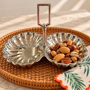 May include: A silver-toned metal double serving dish with a scalloped edge design and a central vertical handle. One side of the dish is empty, while the other side is filled with a variety of mixed unshelled nuts including almonds and pecans.