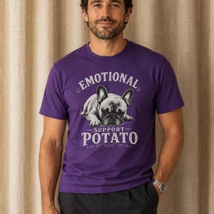 Może przedstawiać: Fioletowa koszulka z napisem "EMOTIONAL SUPPORT - POTATO" i grafiką buldoga francuskiego. Koszulka z kr&oacute;tkim rękawem jest wykonana z miękkiego materiału. Osoba nosi ciemnoszare spodnie i srebrny zegarek.