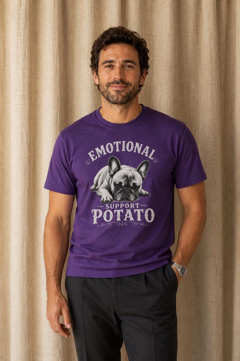 Może przedstawiać: Fioletowa koszulka z napisem "EMOTIONAL SUPPORT - POTATO" i grafiką buldoga francuskiego. Koszulka z kr&oacute;tkim rękawem jest wykonana z miękkiego materiału. Osoba nosi ciemnoszare spodnie i srebrny zegarek.
