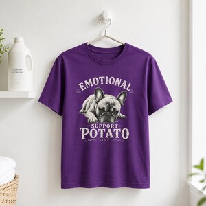 Może przedstawiać: Fioletowa koszulka z napisem "EMOTIONAL SUPPORT POTATO" i grafiką buldoga francuskiego. Koszulka wisi na białej ścianie.