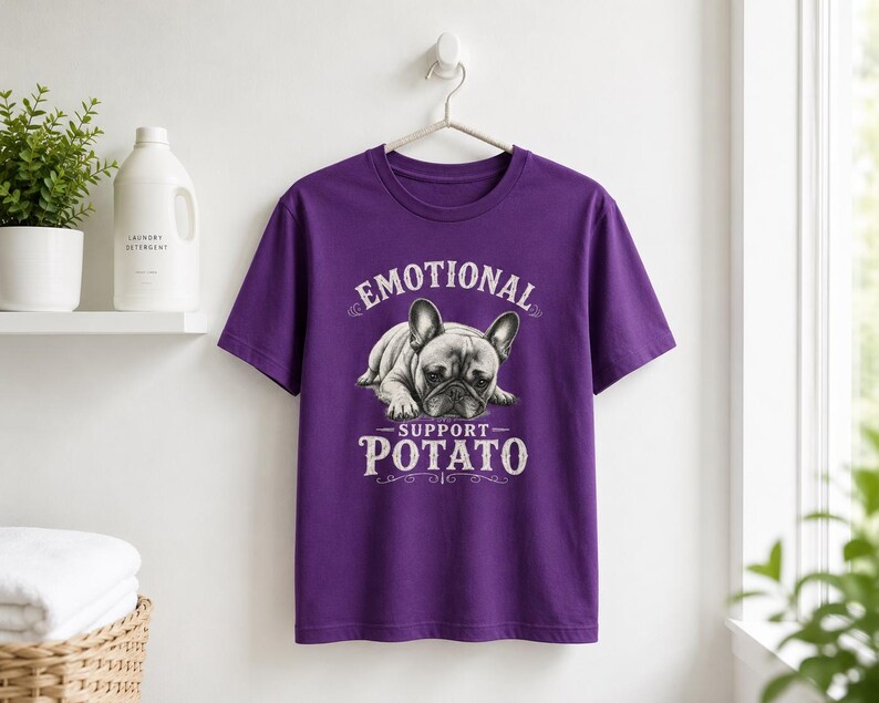 Może przedstawiać: Fioletowa koszulka z napisem "EMOTIONAL SUPPORT POTATO" i grafiką buldoga francuskiego. Koszulka wisi na białej ścianie.