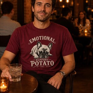 Może przedstawiać: Bordowa koszulka z napisem "EMOTIONAL SUPPORT POTATO" i grafiką buldoga francuskiego. Osoba siedzi przy stole ze szklanką wody i zapaloną świecą. Tło to słabo oświetlona restauracja.