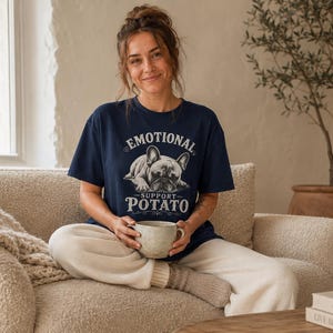 Może przedstawiać: Granatowa koszulka z napisem "EMOTIONAL SUPPORT POTATO" i grafiką buldoga francuskiego. Osoba trzyma beżowy kubek, siedząc na beżowej kanapie. Beżowy koc jest zarzucony na ramię kanapy.